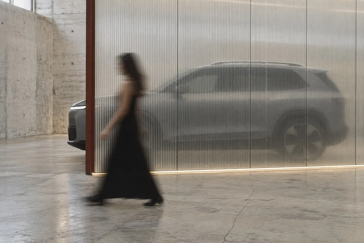 Concept render: a woman walks past a fluted glass pavilion wall, a car silhouette waiting behind it in a pale concrete hall